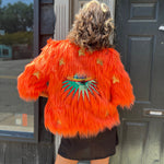 A woman wearing the Planet Queen Orange Faux Fur Jacket with a black dress in front of a window.