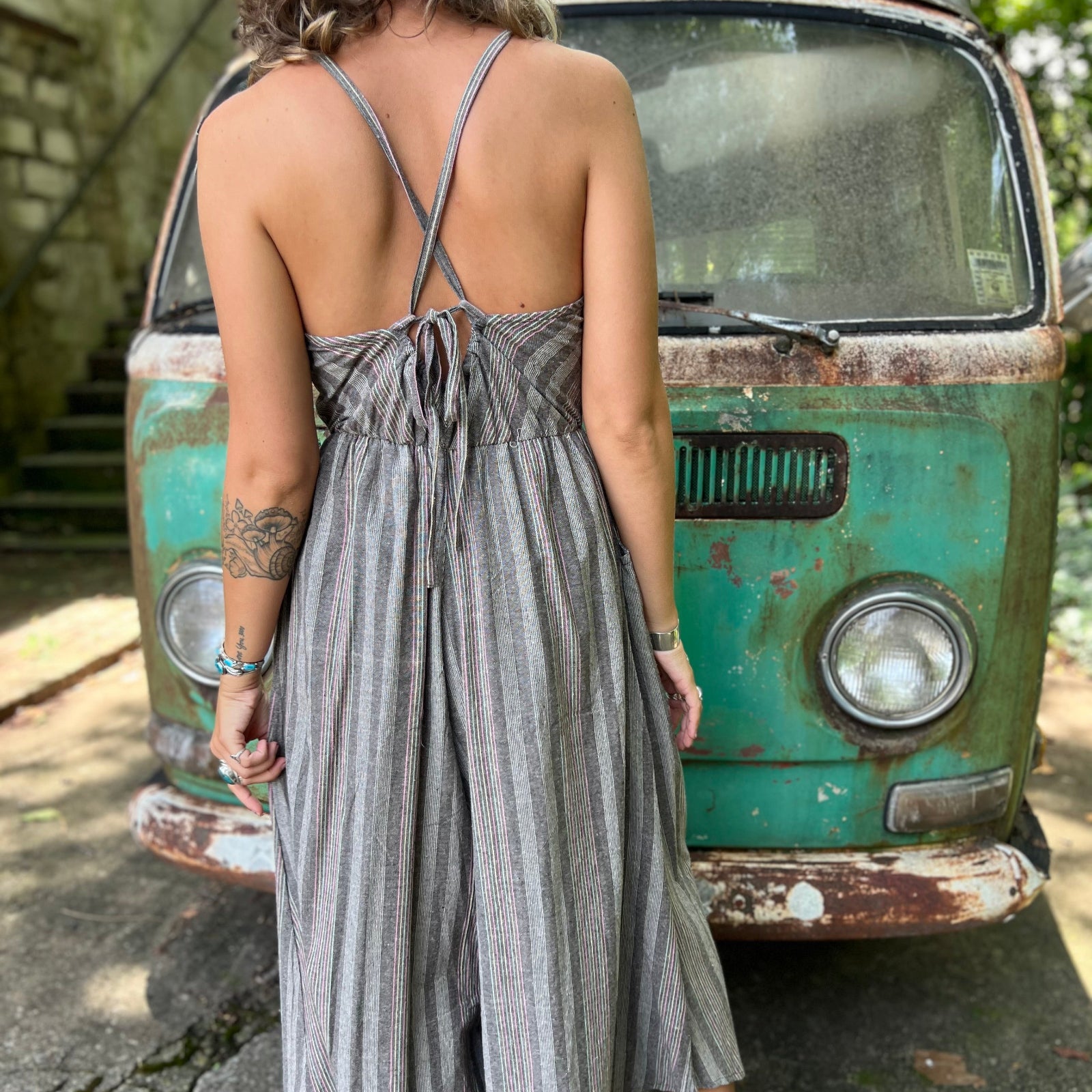 A woman wearing the Grey Striped Palazzo Cotton Jumpsuit in front of a vintage Volkswagen bus, showing off the back.