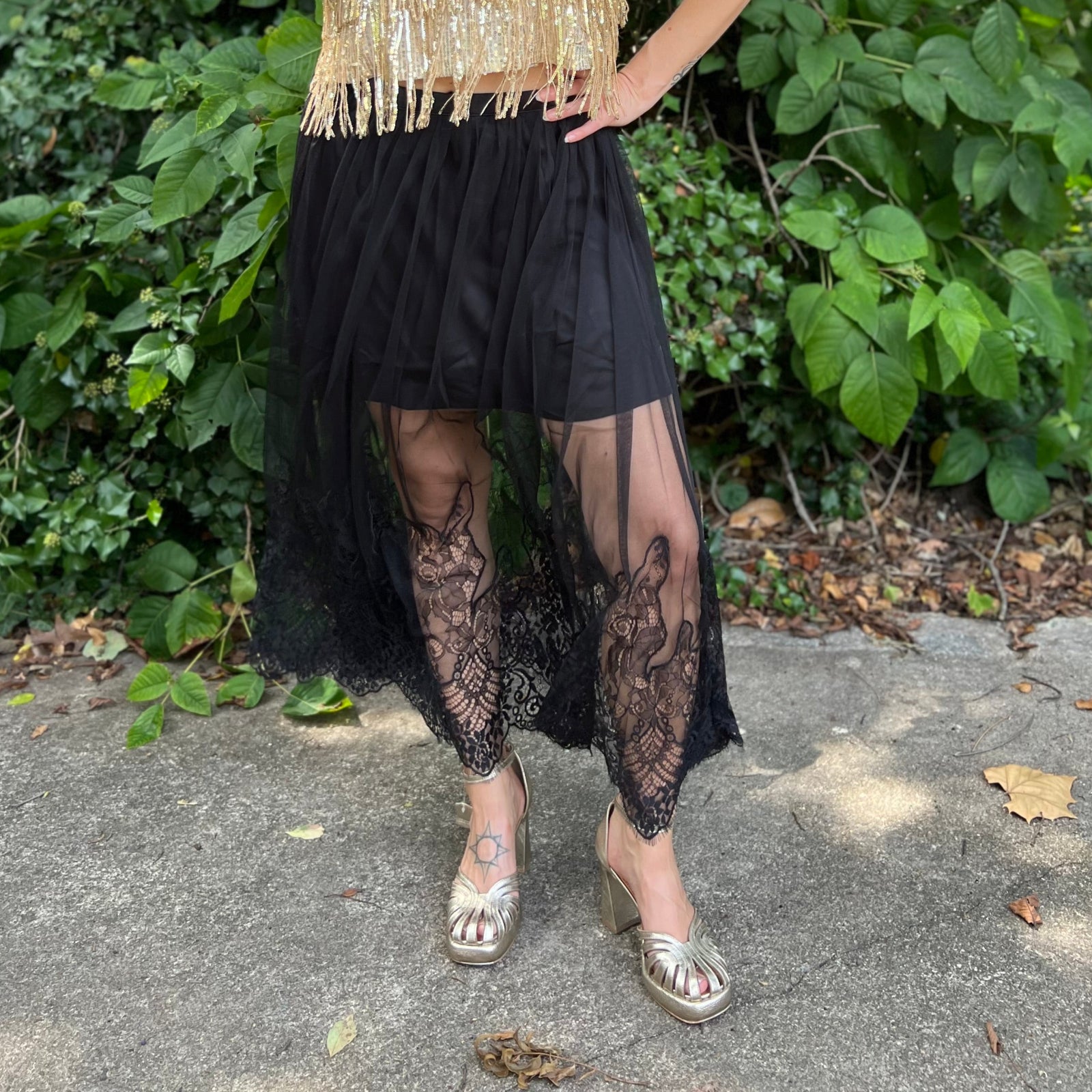 A woman wearing the Sheer Mesh Lace Mini Lined Maxi Skirt with a gold top in front of some greenery.
