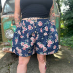 A woman wearing the Floral Print Denim Shorts in front of a teal vintage Volkswagen bus.