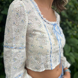 A woman wearing the blue Eyelet Floral Ribbon Trim Cotton Crop Top with jean shorts in front of some greenery.