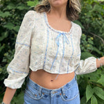 A woman wearing the blue Eyelet Floral Ribbon Trim Cotton Crop Top with jean shorts in front of some greenery.