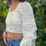 A woman wearing the yellow Eyelet Floral Ribbon Trim Cotton Crop Top with jean shorts in front of some greenery.