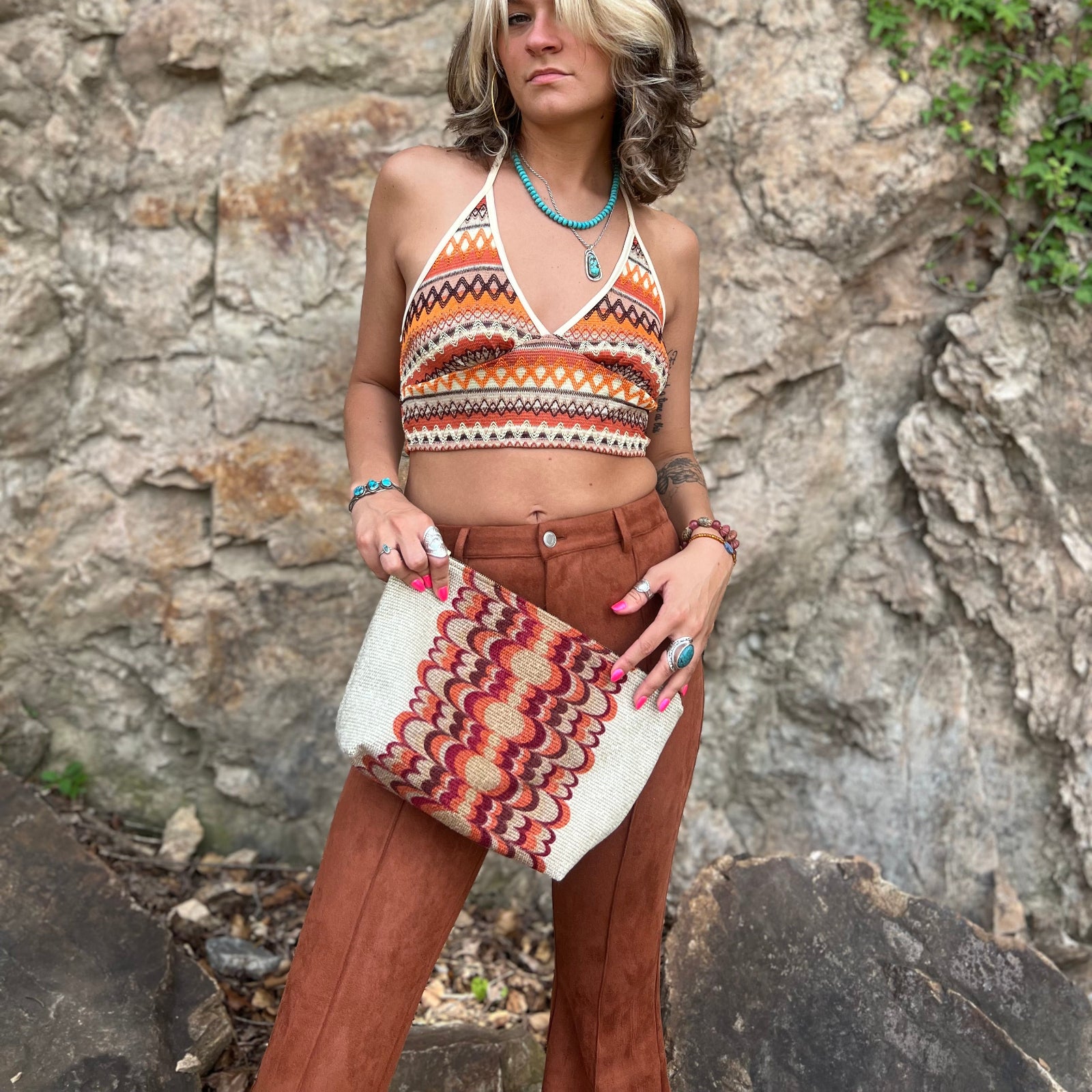 A woman holding the Mid Century Modern Traveller Large Pouch while wearing brown pants and a retro halter top, in front of a stone wall.