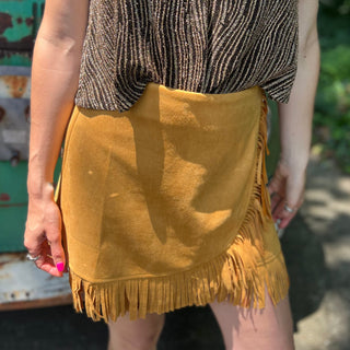 A woman wearing the Mustard Suede Fringe Wrap Skirt with a sparkly top.