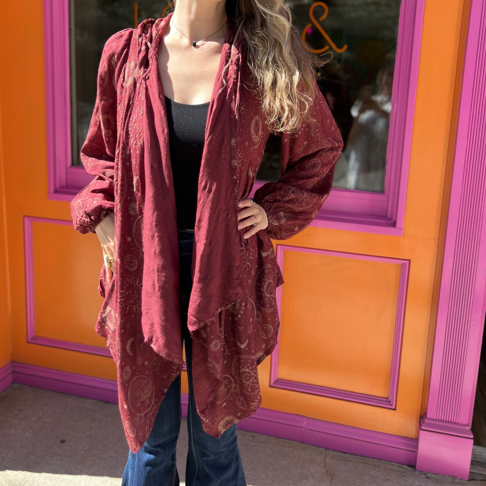 A woman wearing the Flowy Hoodie Waterfall Hooded Cardigan with a black tank and bell bottoms in front of a pink and orange wall.