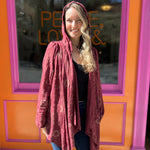 A woman wearing the Flowy Hoodie Waterfall Hooded Cardigan with a black tank and bell bottoms in front of a pink and orange wall.