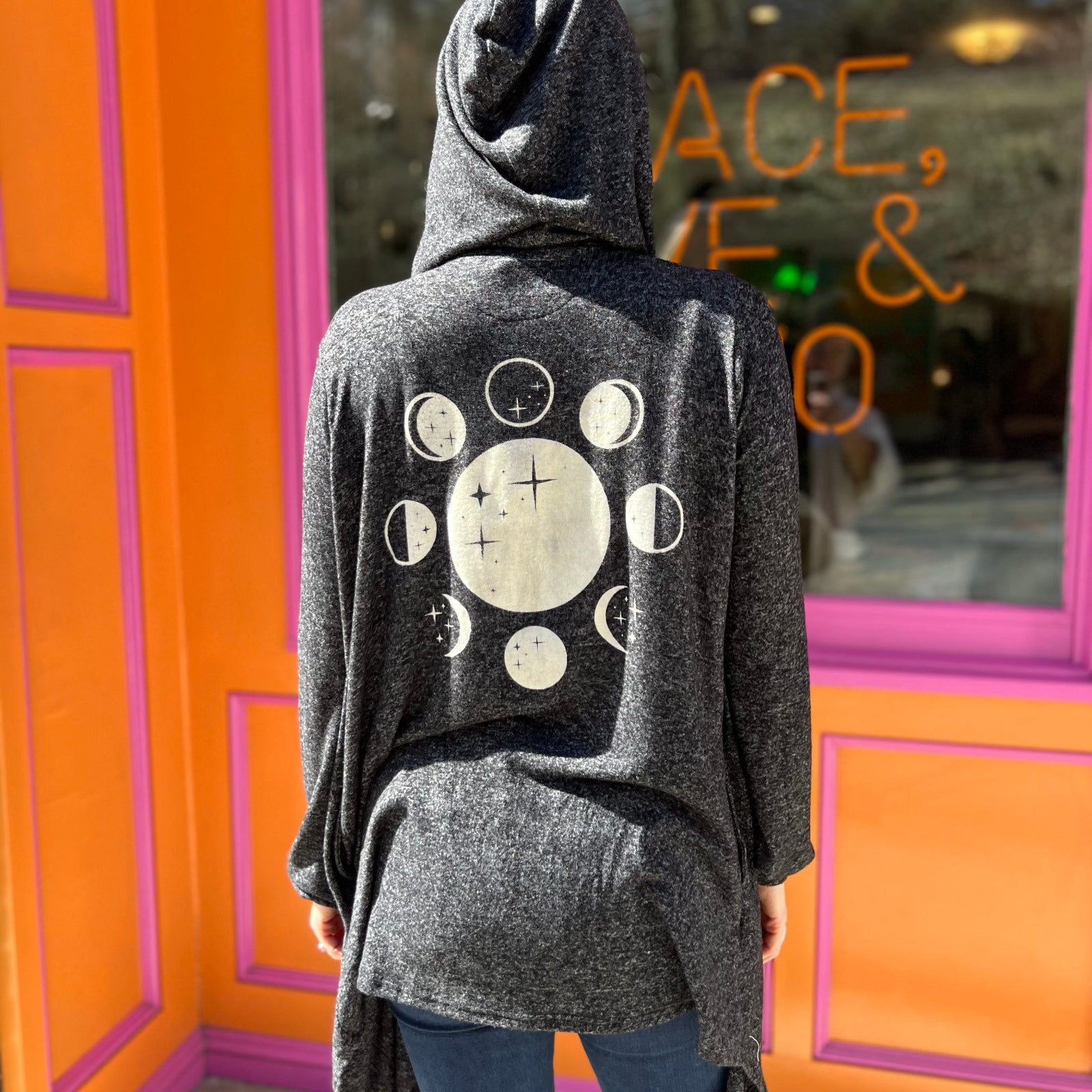 A woman wearing the black Moon Cycle Waterfall Hoodie with a black tank and bellbottoms in front of an orange and pink wall.