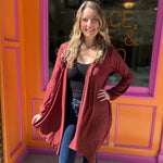 A woman wearing the maroon Moon Cycle Waterfall Hoodie with a black tank and bellbottoms in front of an orange and pink wall.