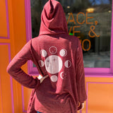 A woman wearing the maroon Moon Cycle Waterfall Hoodie with a black tank and bellbottoms in front of an orange and pink wall.