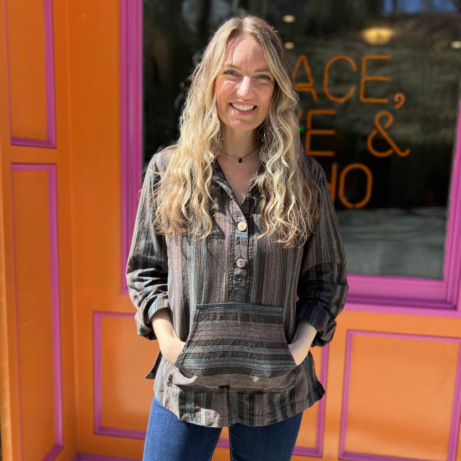 A woman wearing the agate Womens Half Button Baja Hoodie with jeans in front of a pink and orange wall.
