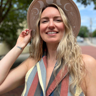 Close-up of woman smiling in celestial phase hat and retro-striped top, with “Let it Be” tattoo on wrist