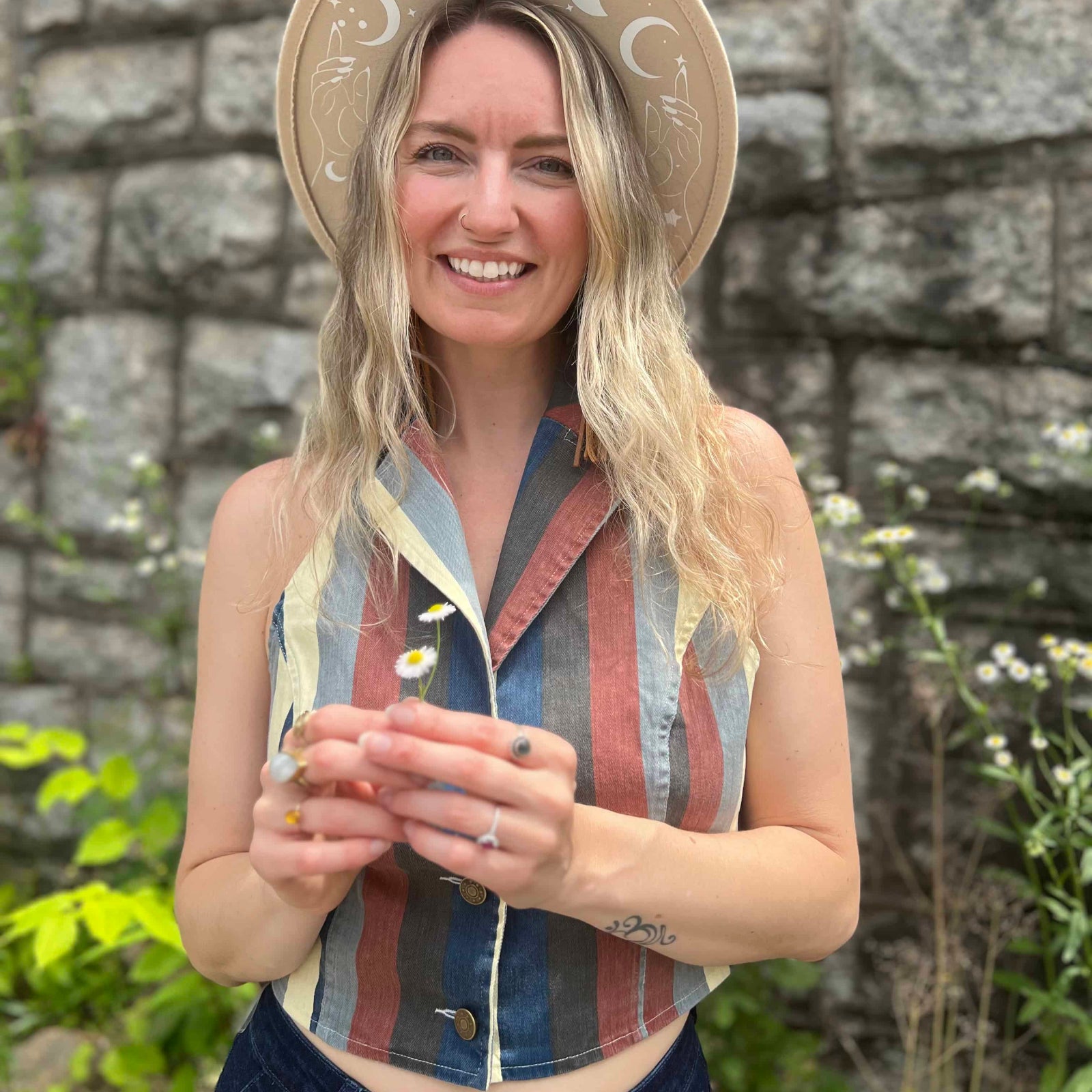 Woman in boho moon hat holding daisies, wearing a striped halter crop top and high-waisted jeans