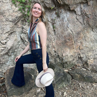 Woman wearing vintage-inspired striped top and bell-bottom jeans standing on a rock with hat in hand