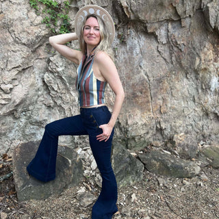 Woman in striped vest top and flared jeans striking a confident pose with leg on rock and hand on hat