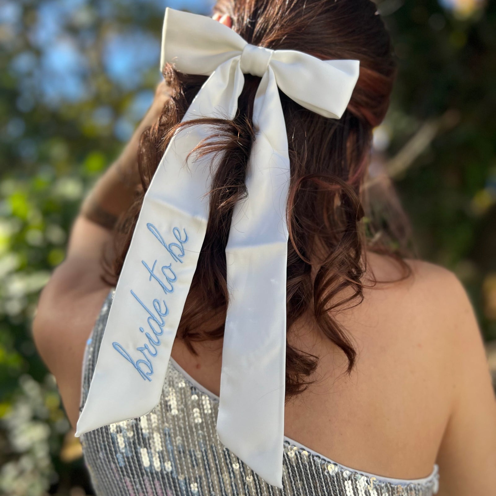A woman wearing the Bride To Be Embroidered White Satin Bow with a silver sequin dress.