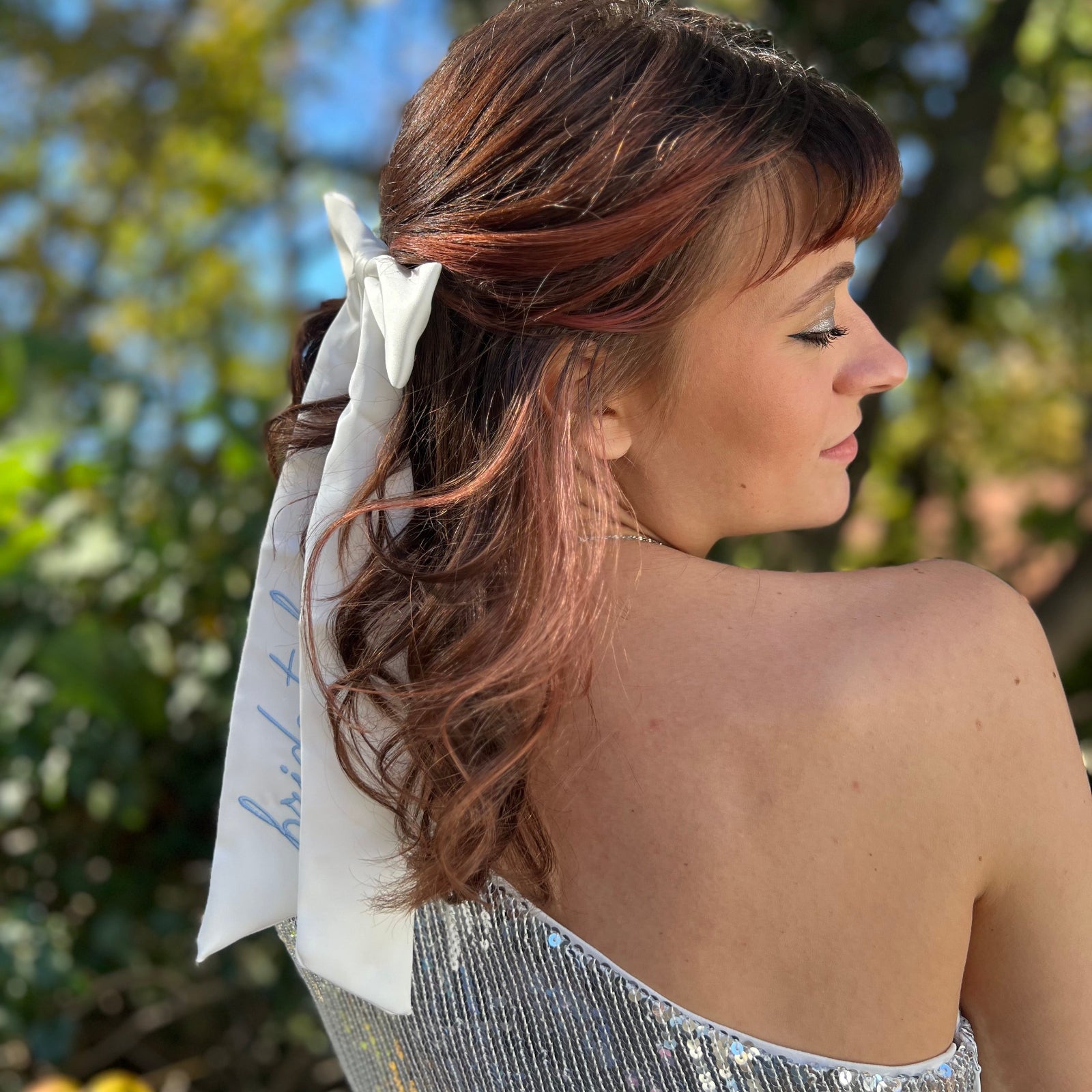 A woman wearing the Bride To Be Embroidered White Satin Bow with a silver sequin dress, showing off the side.