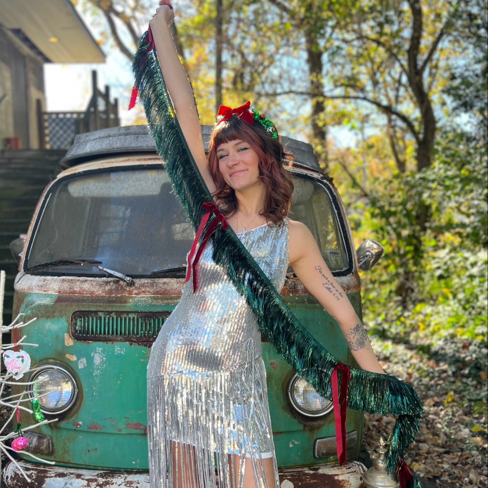 A woman in a silver sequin dress holding the Xmas Velvet Bow & Green Fringe Banner in front of a vintage teal Volkswagen bus.