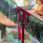 An up-close image of a woman in a silver sequin dress holding the Xmas Velvet Bow & Green Fringe Banner in front of a vintage teal Volkswagen bus.