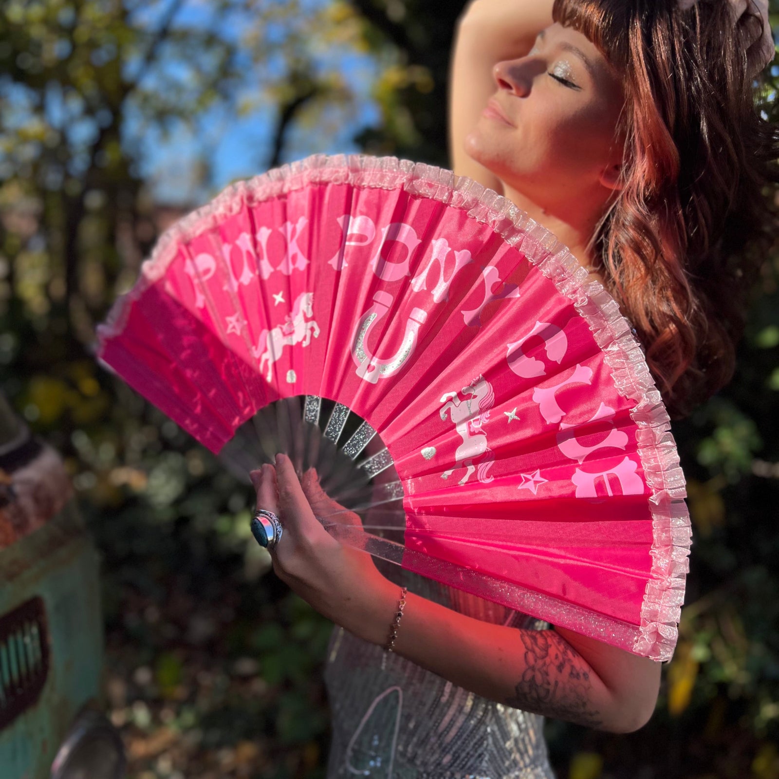 A woman in a silver sequin dress holding the Pink Pony Club Ruffled Folding Fan in front of herself.