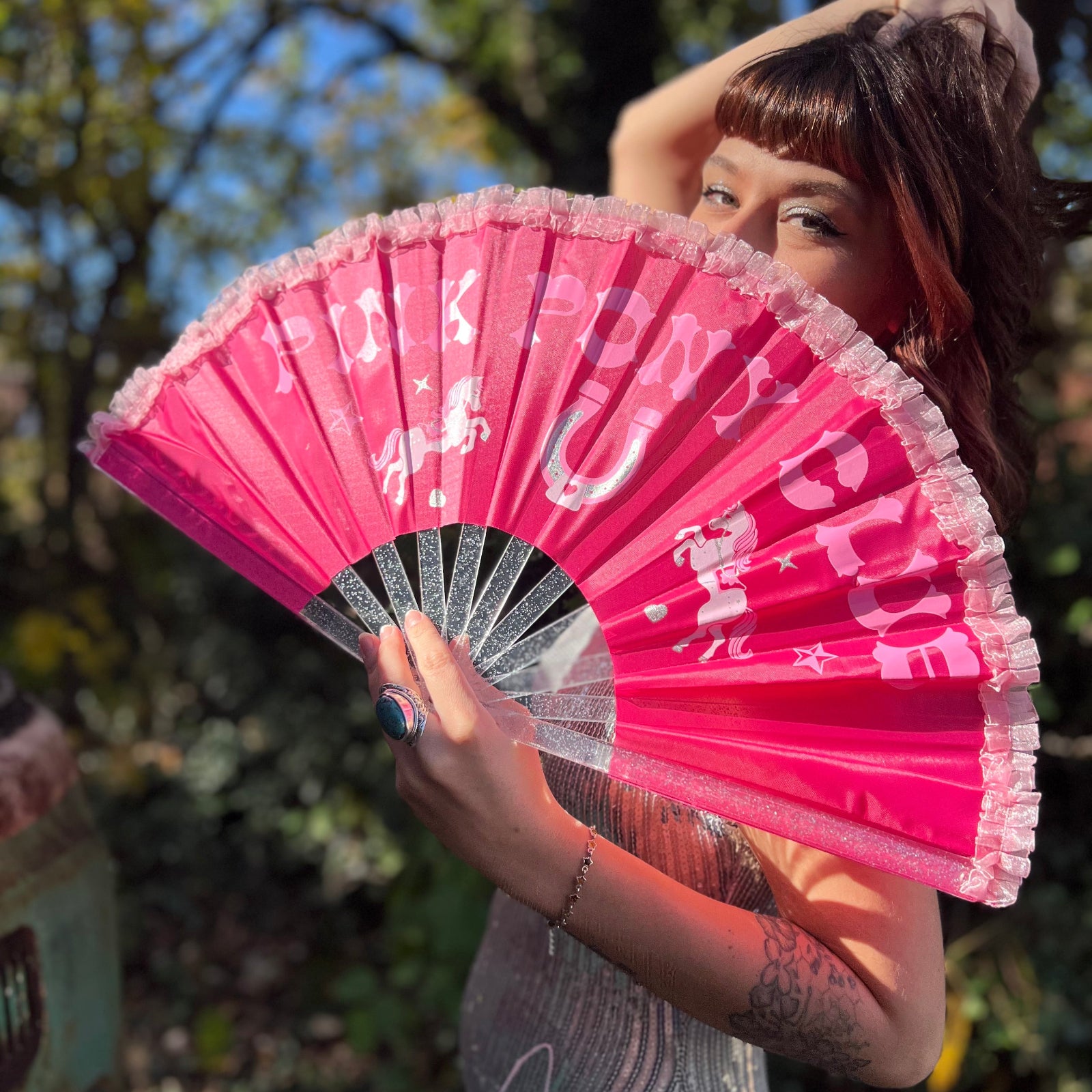 A woman in a silver sequin dress holding the Pink Pony Club Ruffled Folding Fan in front of her face.
