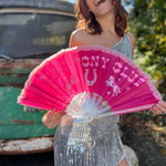 A woman in a silver sequin dress holding the Pink Pony Club Ruffled Folding Fan in front of herself.