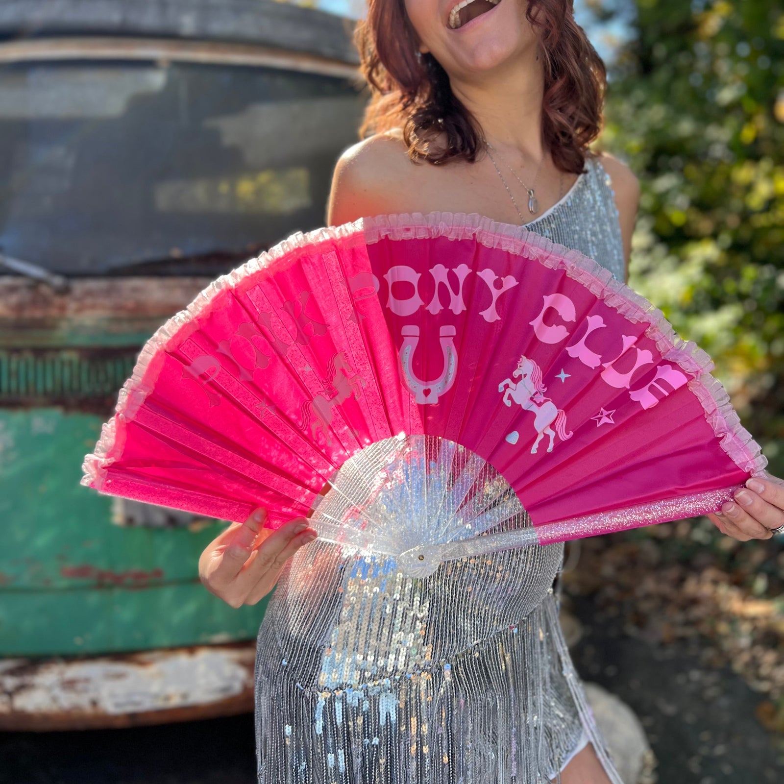 A woman in a silver sequin dress holding the Pink Pony Club Ruffled Folding Fan in front of herself.