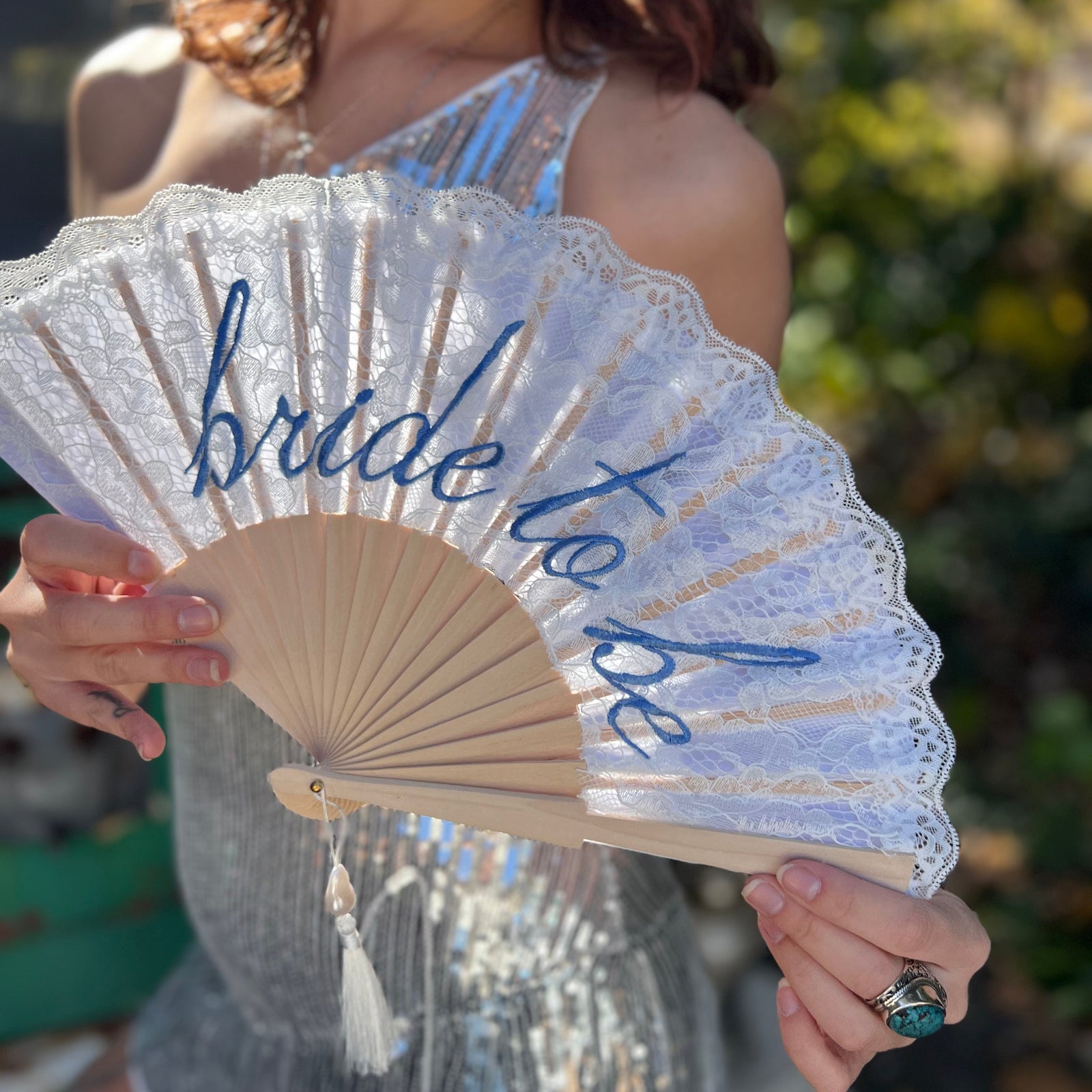 A woman in a silver sequin dress holding a Bride to Be Lace Embroidered Folding Fan.