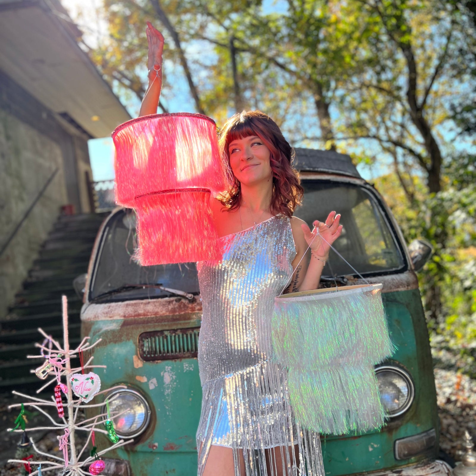 A woman in a silver dress holding two Fringe Party Chandeliers in front of a vintage teal Volkswagen bus.