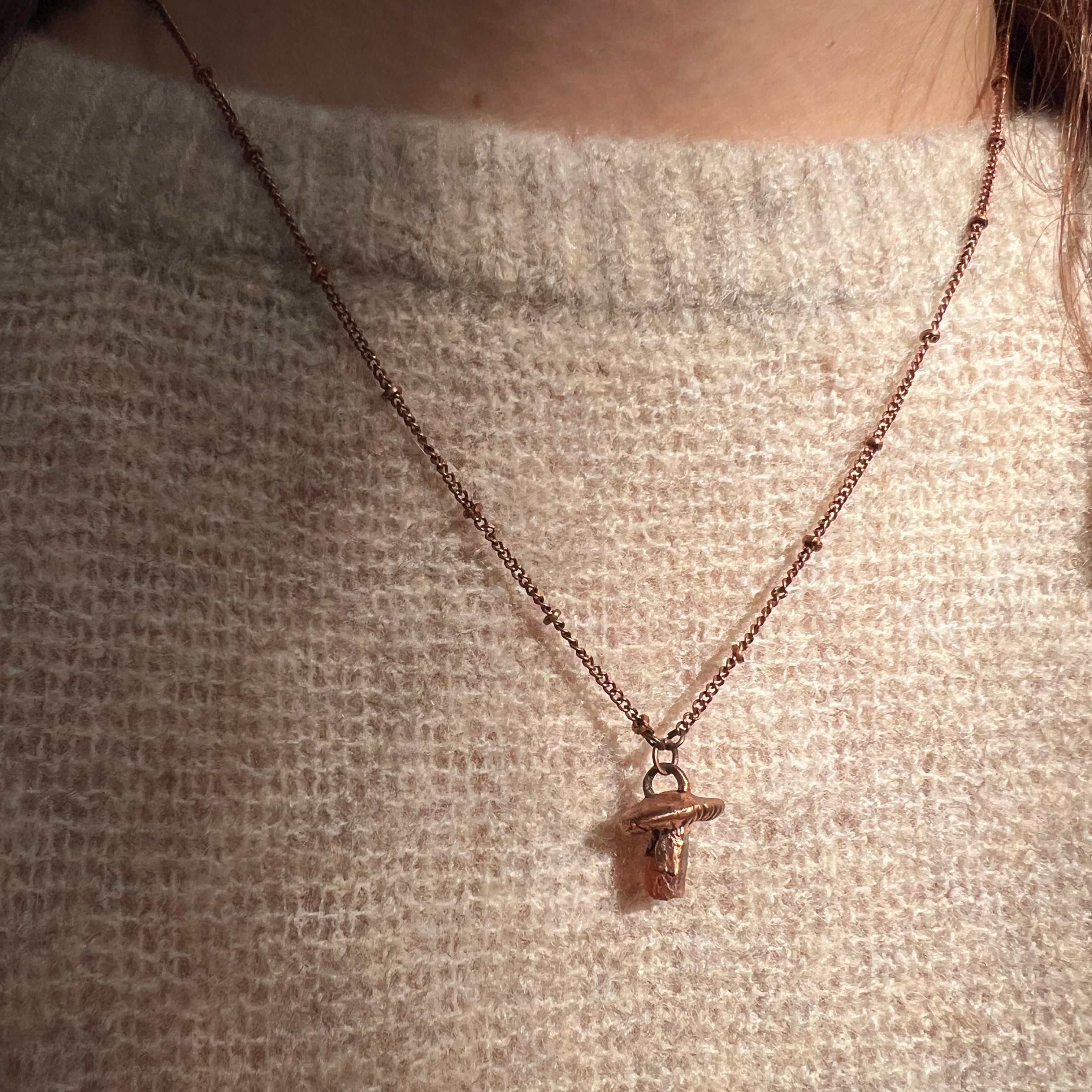 Someone wearing the Dainty Orange Kyanite Crystal Copper Mushroom Necklace with a beige sweater.