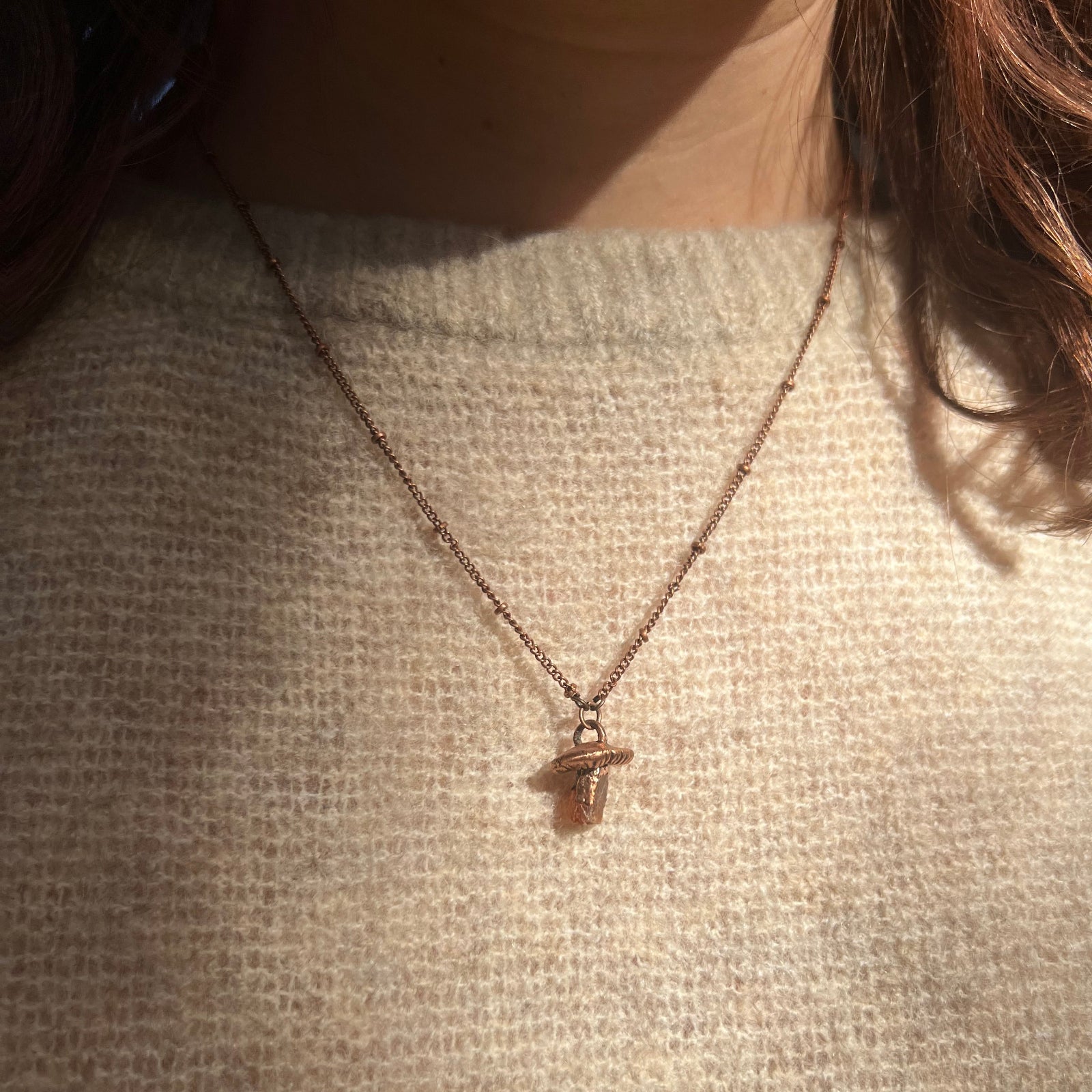Someone wearing the Dainty Orange Kyanite Crystal Copper Mushroom Necklace with a beige sweater.