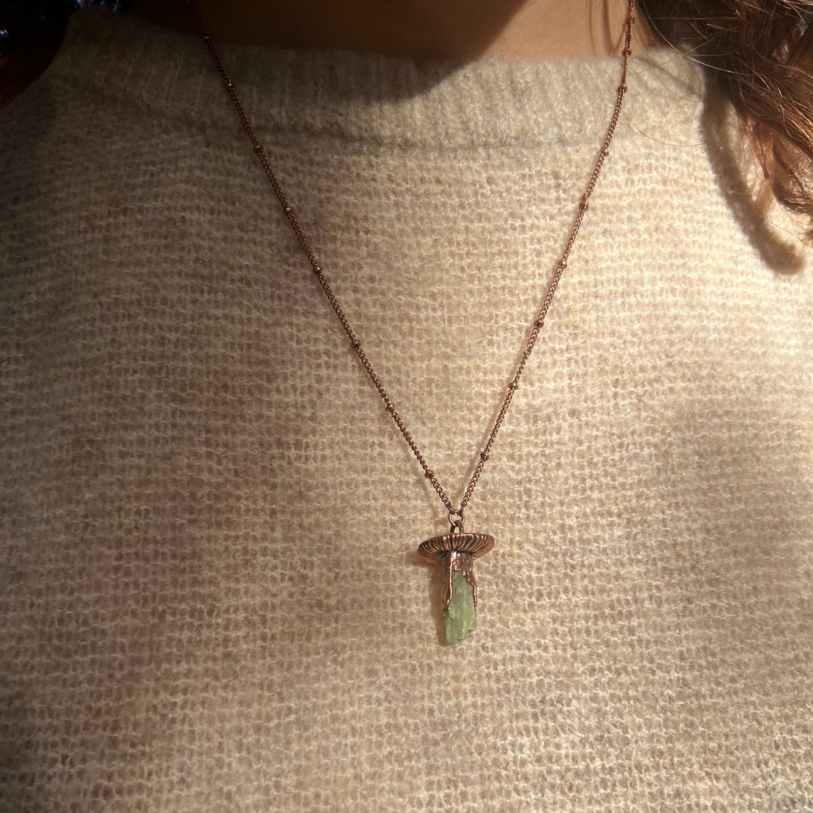Someone wearing the Dainty Green Kyanite Crystal Copper Mushroom Necklace with a beige sweater.
