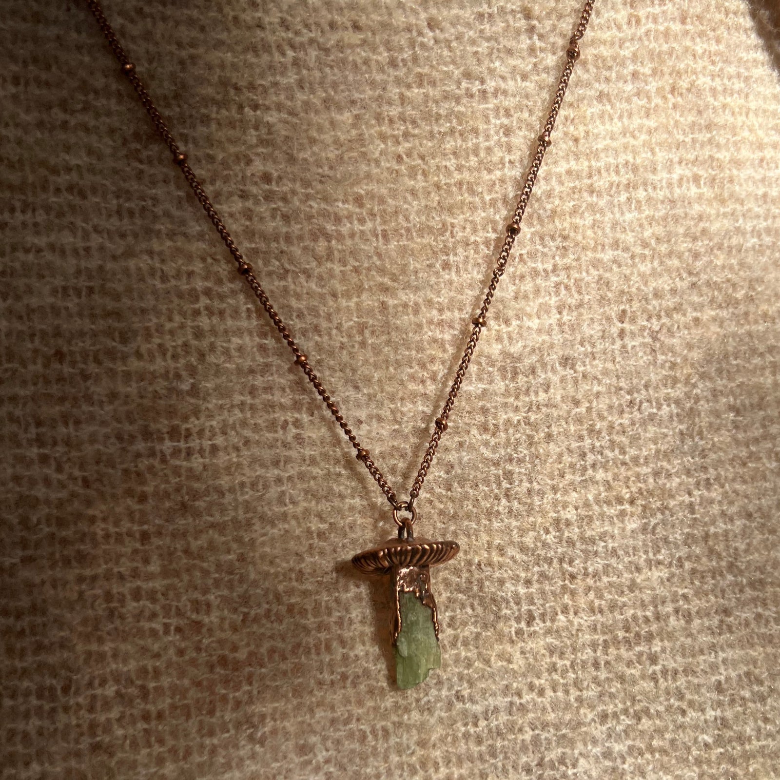 Someone wearing the Dainty Green Kyanite Crystal Copper Mushroom Necklace with a beige sweater.
