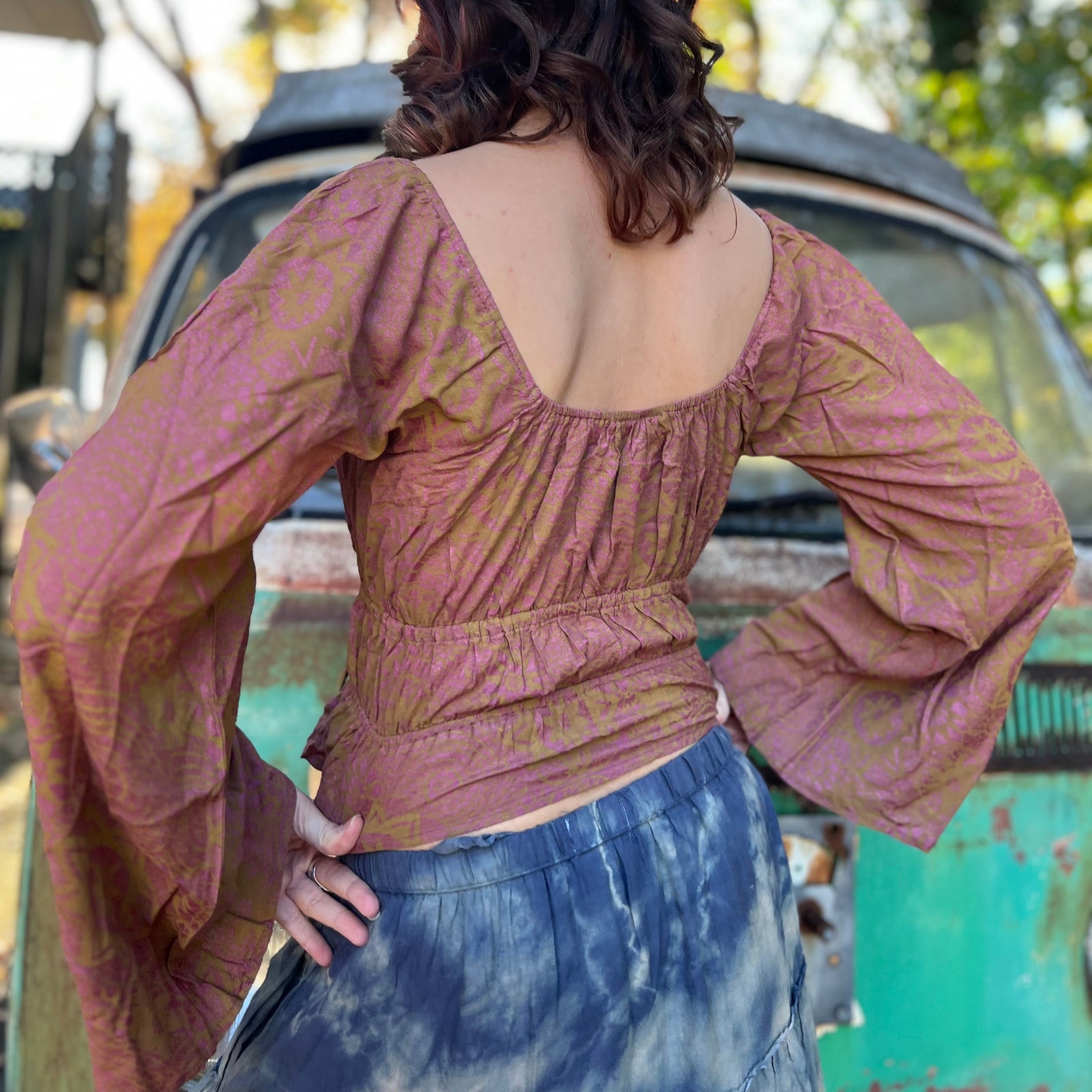 A woman wearing the maroon Flowy Peasant Viscose Print Blouse with a skirt in front of the vintage teal Volkswagen bus, showing off the back.