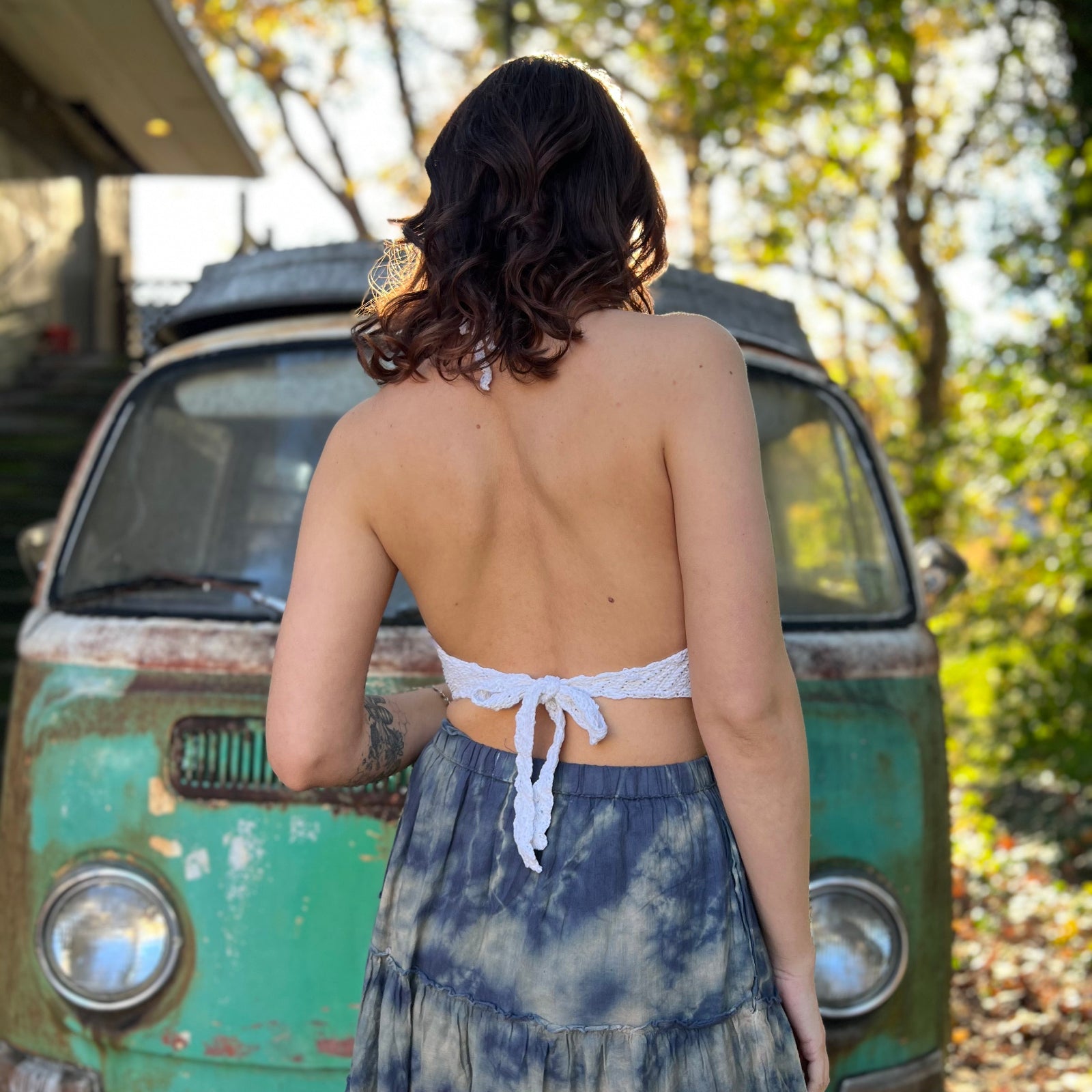 A woman wearing the Moon Cycle Crochet Halter Cotton Top with a skirt in front of a vintage teal Volkswagen bus, showing off the back.
