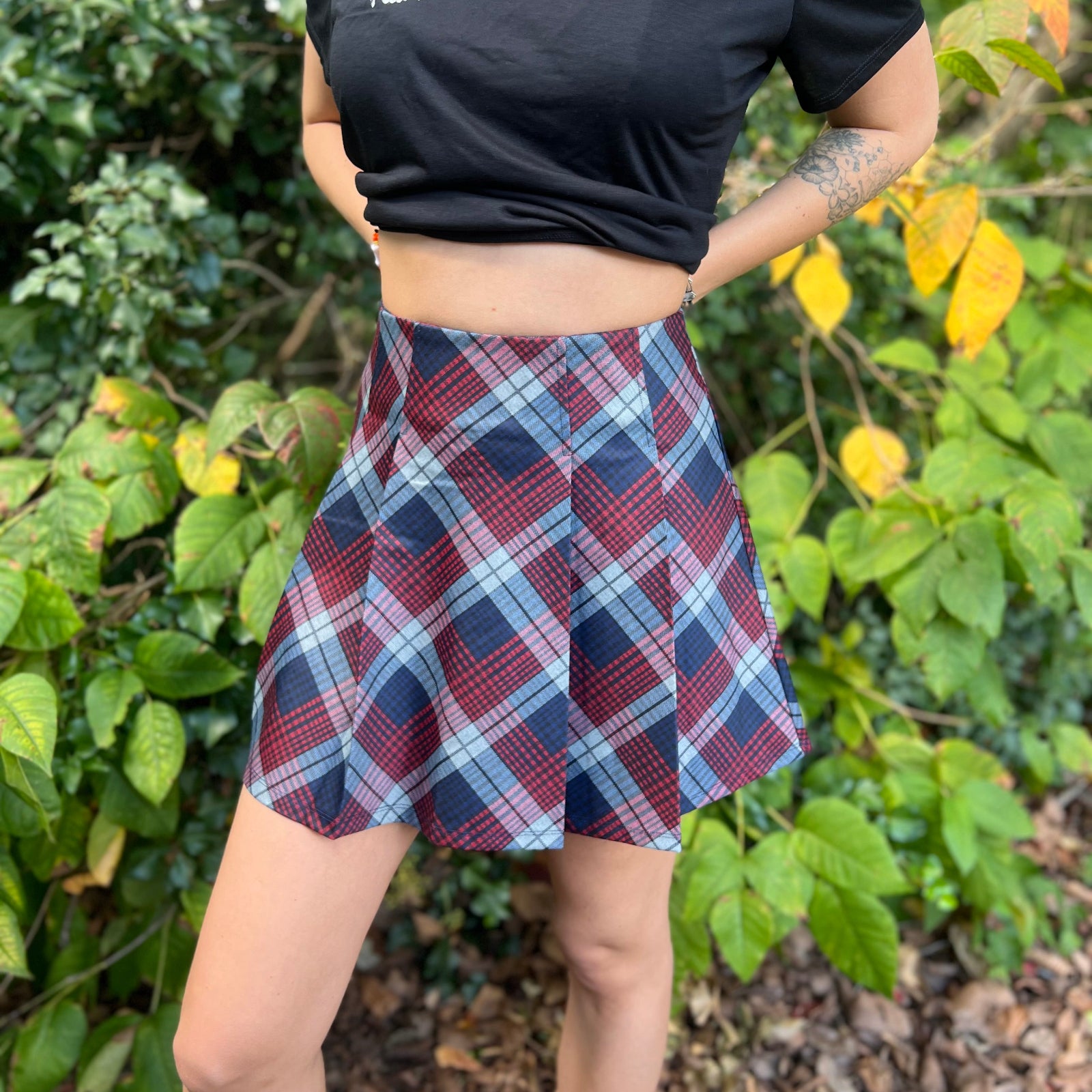 A woman wearing the Plaid Pleated Mini Skirt with a black shirt in front of greenery.