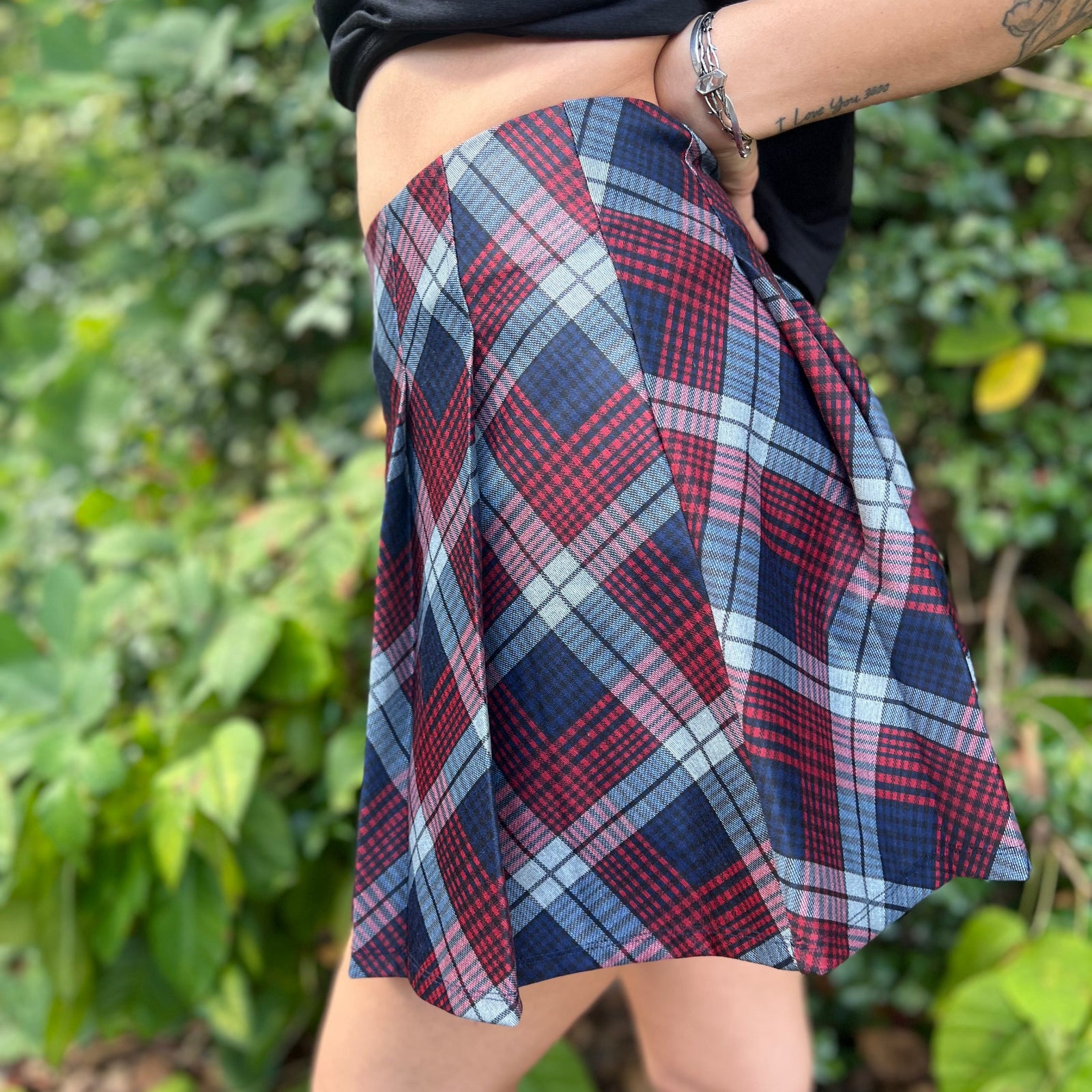A woman wearing the Plaid Pleated Mini Skirt with a black shirt in front of greenery.