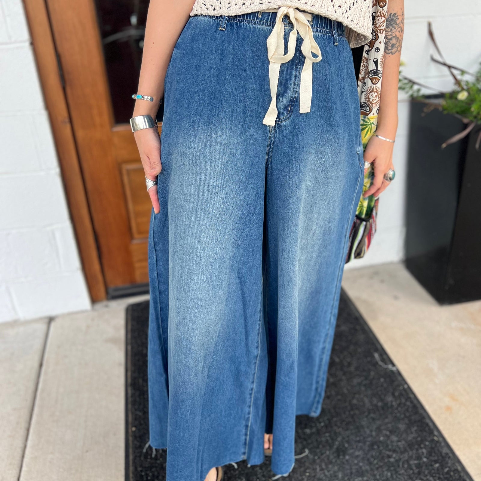 A woman wearing the dark wash Denim Drawstring Waist Band Wide Leg Pants with a cream crocheted top.