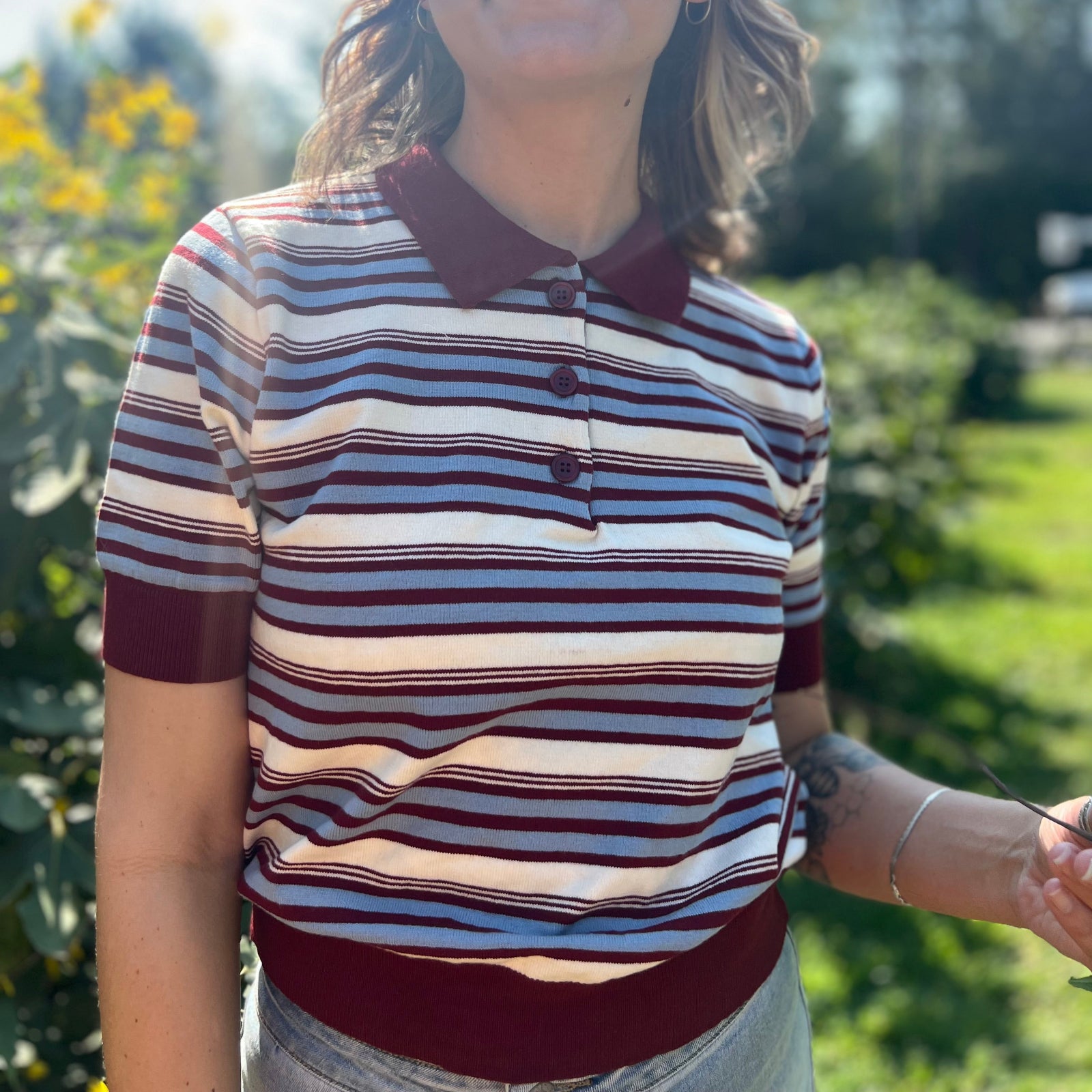 A woman wearing the Striped Knit Sweater Half Button Up Collar Top with some jeans on, in front of some greenery.