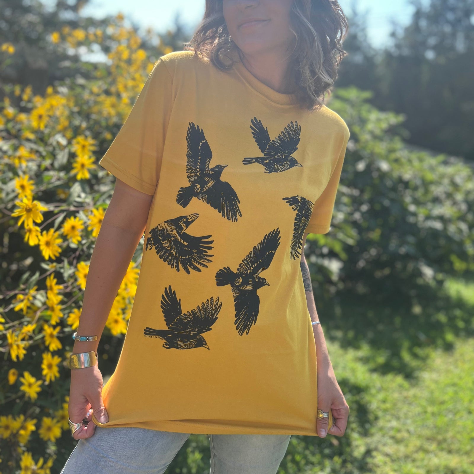 A woman wearing the Crows Organic Cotton T Shirt with some jeans in front of some greenery.
