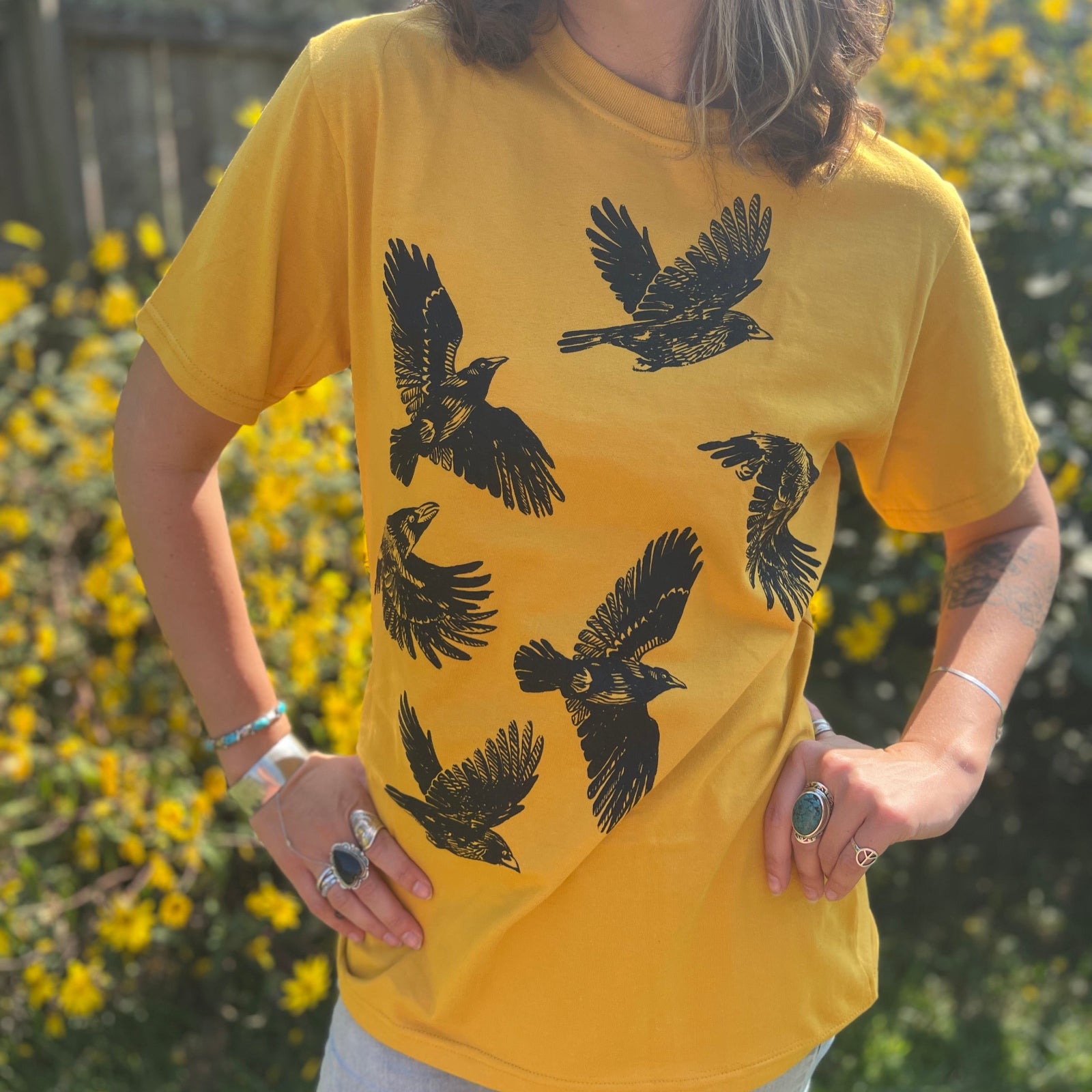 A woman wearing the Crows Organic Cotton T Shirt with some jeans in front of some greenery.