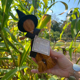 Someone holding a Witch Samhain Folk Corn Husk Poppet Doll in front of greenery.