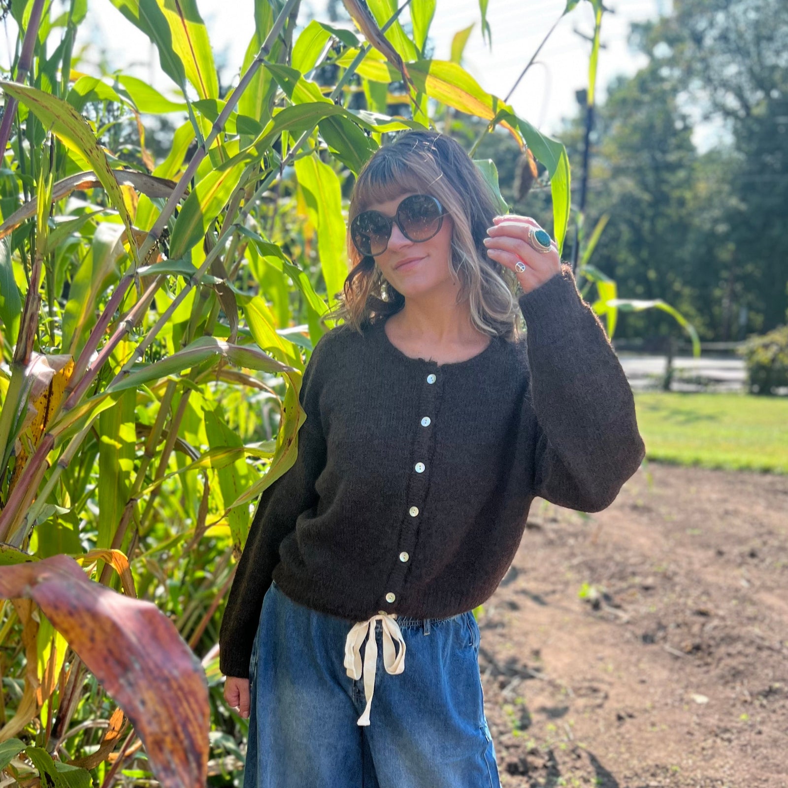 A woman wearing the dark chocolate Knit Round Neck Sweater Cardigan Top with jeans in front of some greenery.