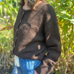 A woman wearing the dark chocolate Knit Round Neck Sweater Cardigan Top with jeans in front of some greenery.