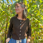A woman wearing the dark chocolate Knit Round Neck Sweater Cardigan Top with jeans in front of some greenery.