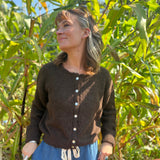 A woman wearing the dark chocolate Knit Round Neck Sweater Cardigan Top with jeans in front of some greenery.