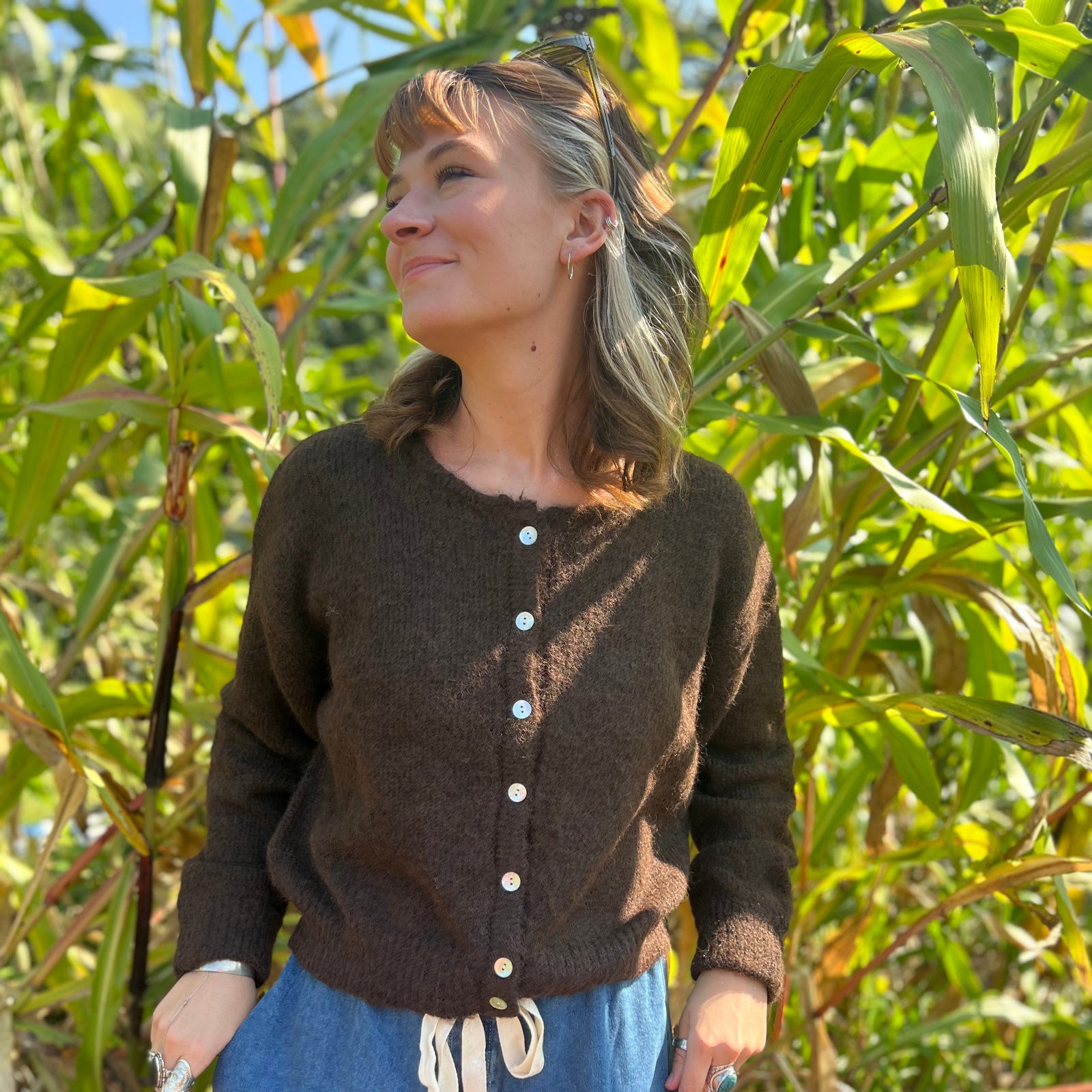 A woman wearing the dark chocolate Knit Round Neck Sweater Cardigan Top with jeans in front of some greenery.