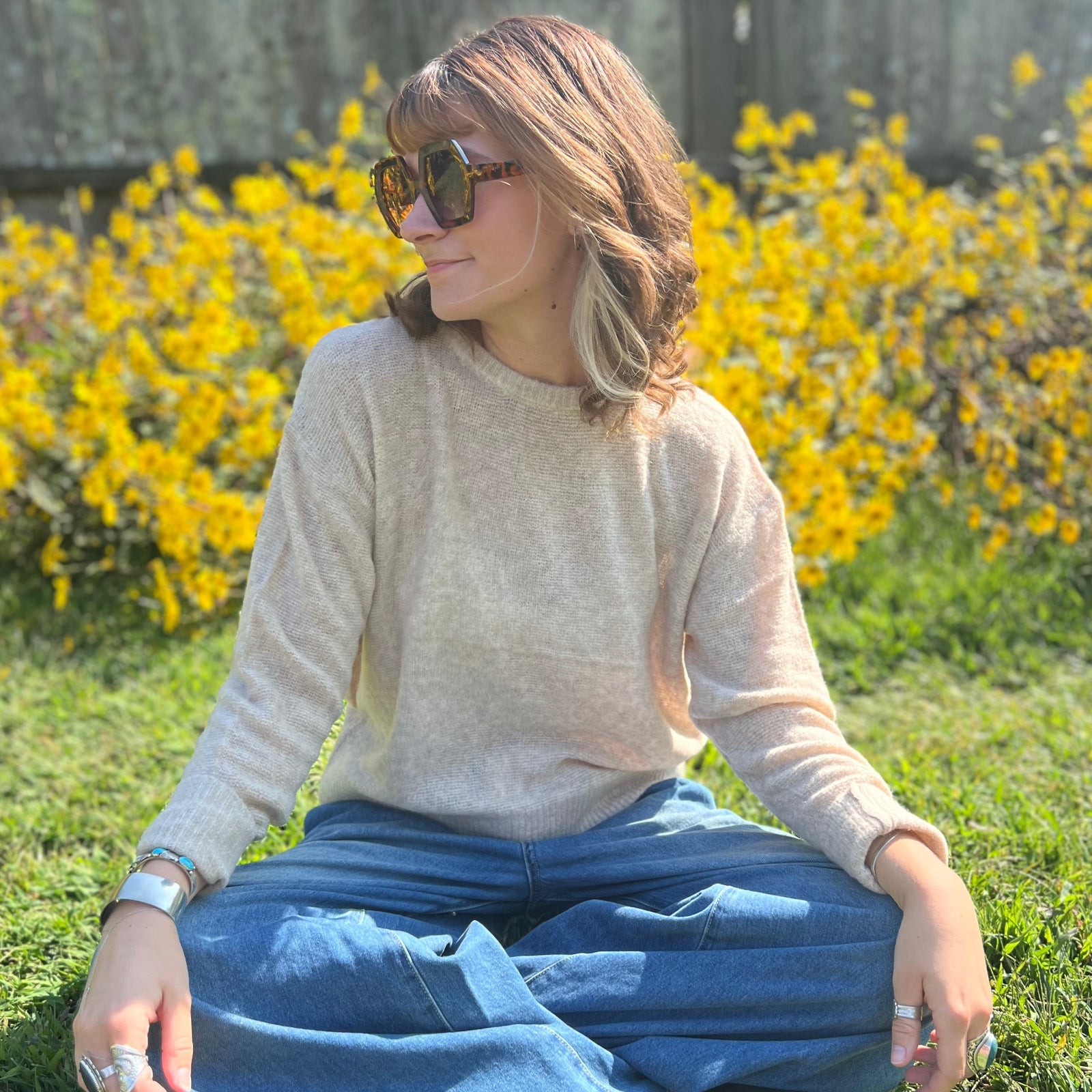 A woman sitting down and wearing the Beige Knit Nylon Wool Spandex Sweater with jeans in front of yellow flowers.