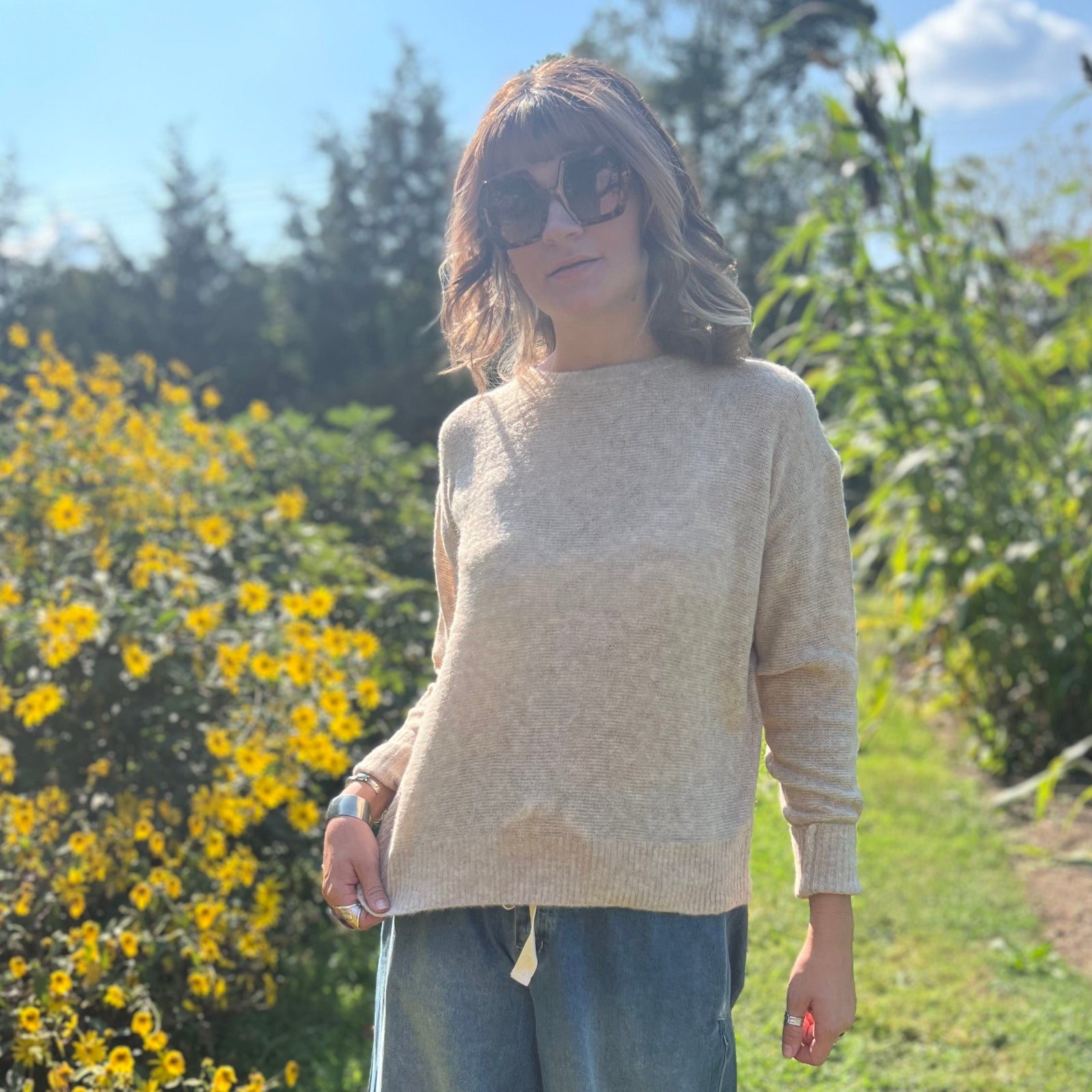 A woman wearing the Beige Knit Nylon Wool Spandex Sweater with jeans in front of yellow flowers.