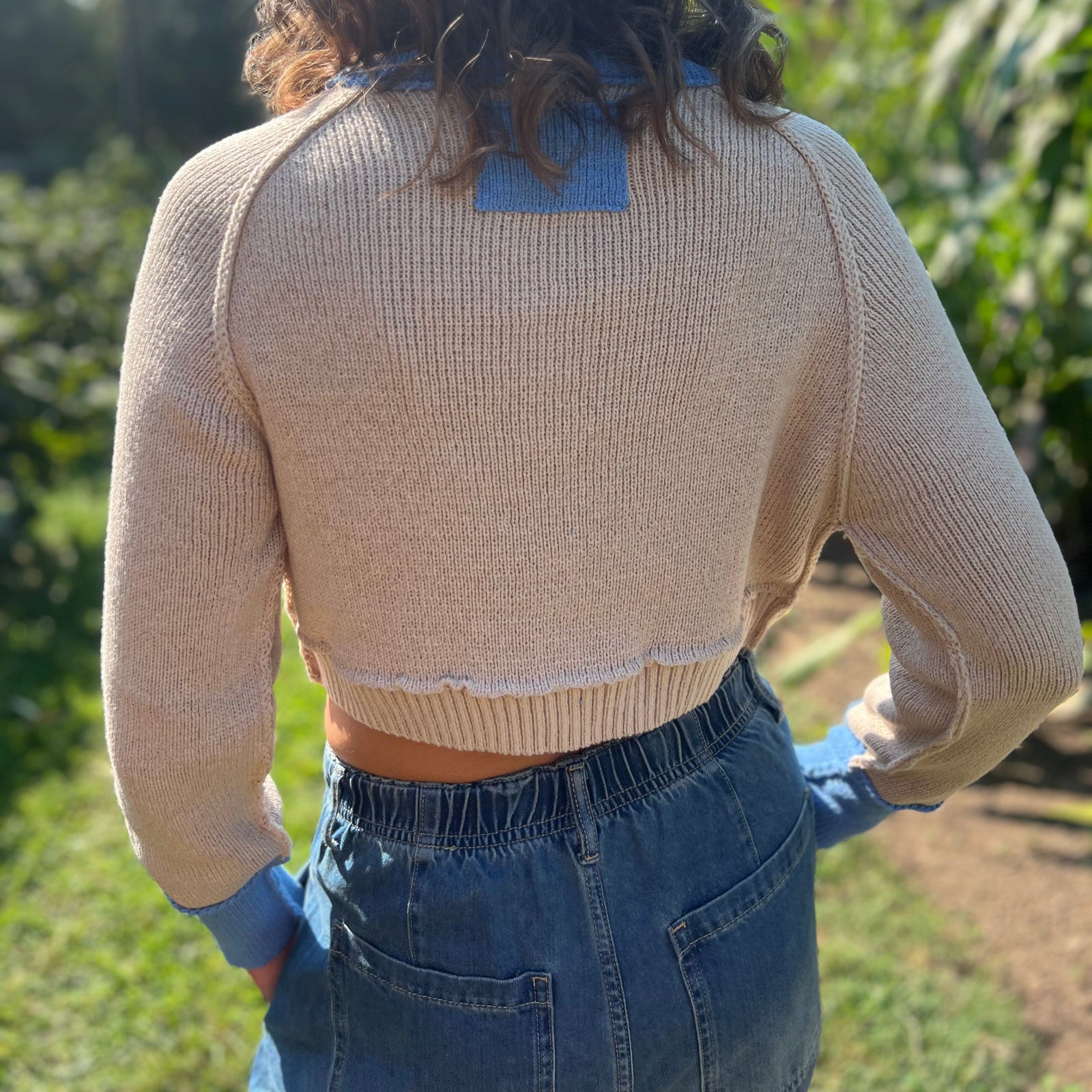 A woman wearing the Contrast Knitted Crew Neck Button Down Cardigan Sweater with denim pants in front of greenery, showing off the back.